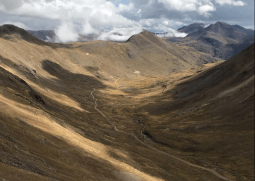 A scenic valley with rolling hills, patches of sunlight, and clouds overhead, showcasing a winding path through the landscape.