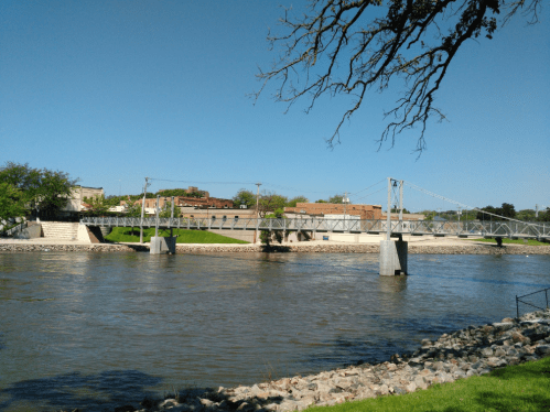 A scenic view of a river with a suspension bridge and green park area under a clear blue sky.