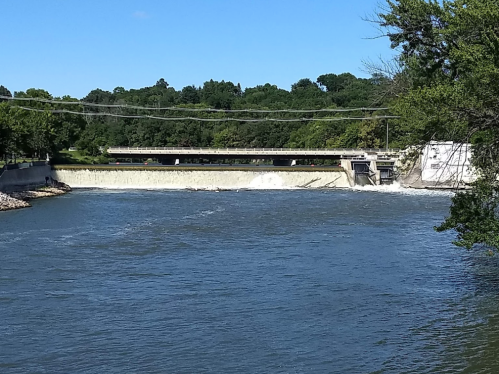 A river flows past a dam with a spillway, surrounded by green trees and a clear blue sky.