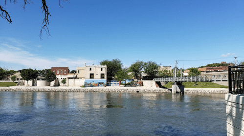 A serene river scene with a clear blue sky, featuring buildings and a mural along the riverbank.