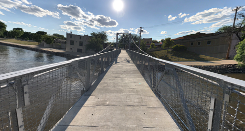 A pedestrian bridge spans a river, with sunlight shining above and buildings visible along the riverbank.