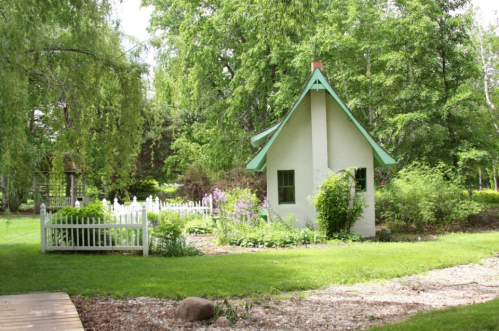 A small, charming cottage with a green roof surrounded by lush greenery and a white picket fence in a serene garden.