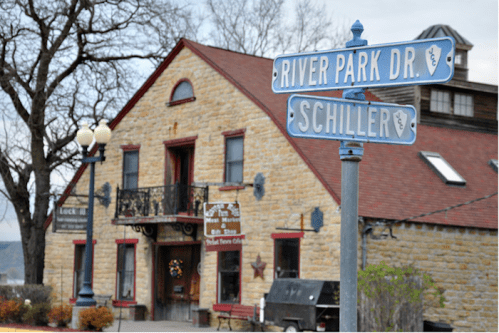 A stone building with a red roof at the intersection of River Park Dr. and Schiller, featuring decorative signs.