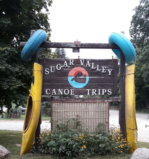 Sign for Sugar Valley Canoe Trips, featuring colorful kayaks and surrounded by greenery and flowers.