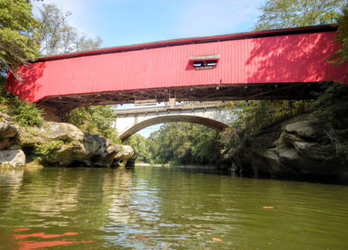 A red covered bridge spans over a river, with a stone arch bridge visible above it, surrounded by lush greenery.