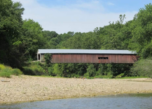A red covered bridge spans a river, surrounded by lush green trees and a sandy riverbank under a blue sky.
