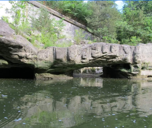 A rocky arch over calm water, surrounded by lush greenery and a stone wall in the background.