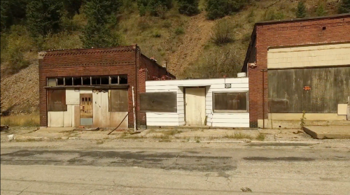 Abandoned buildings with boarded windows, set against a hillside. The scene conveys a sense of neglect and decay.