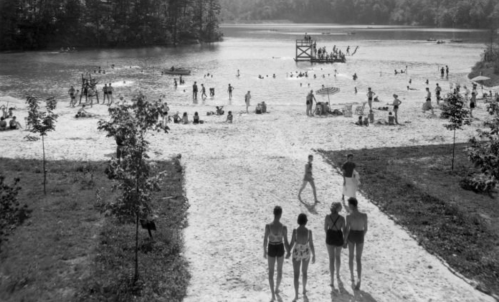 A black-and-white photo of a busy beach with people swimming, sunbathing, and a lifeguard tower in the background.