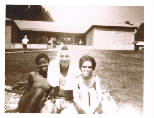 A black-and-white photo of three people sitting on grass, smiling, with a building in the background.