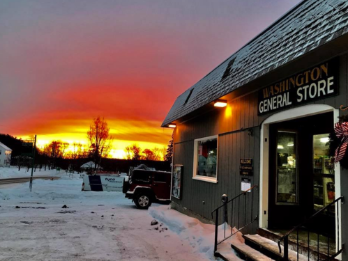 A general store at sunset, with vibrant orange and purple skies, surrounded by snow-covered ground and trees.