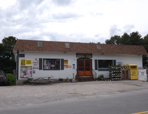 A small, white general store with a brown shingled roof, featuring a wooden door and various signs outside.