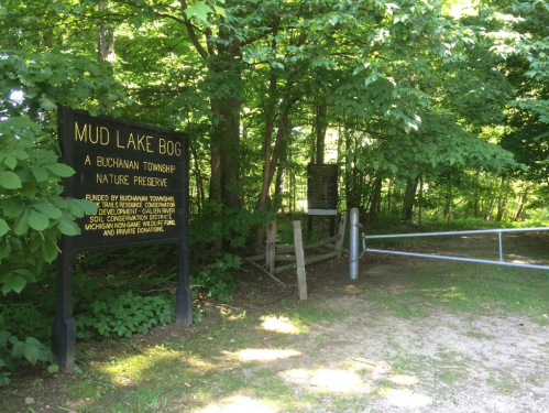 Sign for Mud Lake Bog Nature Preserve, surrounded by trees, with a gated entrance and gravel path.