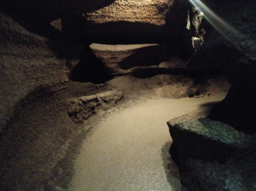A dimly lit cave interior with rocky walls and a sandy floor, featuring uneven surfaces and ledges.