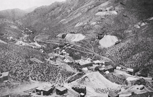 Black and white image of a mining town nestled in a mountainous landscape, with buildings and mining structures visible.