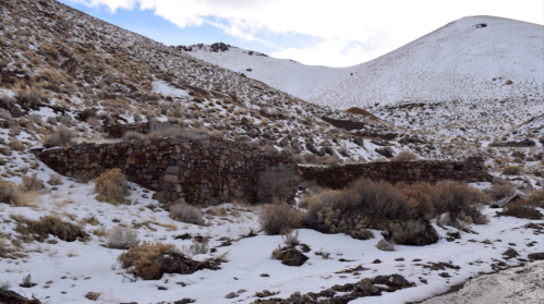 Ruins of a stone structure on a snowy hillside, surrounded by sparse vegetation and rocky terrain.