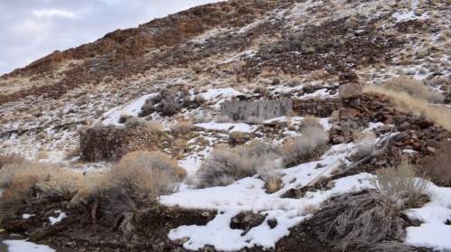 Ruins of stone structures on a snowy hillside, surrounded by dry grass and rocky terrain.