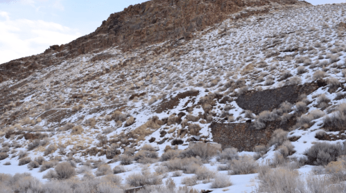A rocky hillside covered in snow and sparse vegetation under a cloudy sky.