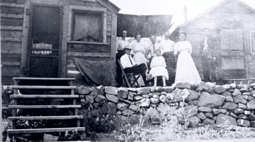 A black and white photo of a group of people posing on a porch in front of a rustic wooden house.