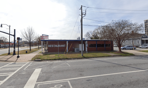 A brick restaurant building on a street corner, with a sign and trees nearby under a cloudy sky.