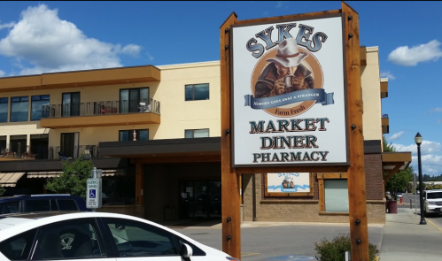 Sign for Sykes Market Diner Pharmacy, featuring a cowboy graphic, with a building in the background and blue sky.