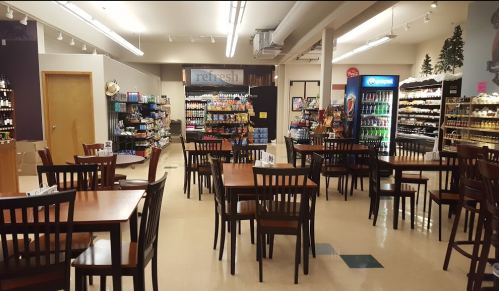 A spacious café with wooden tables and chairs, shelves of snacks, and beverage coolers in the background.