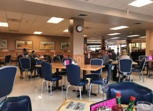 A cozy diner interior with wooden tables, blue chairs, and patrons enjoying meals. Wall art and a clock are visible.