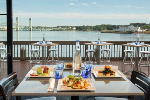 A scenic waterfront restaurant table set with gourmet dishes and drinks, overlooking a bridge and calm water.