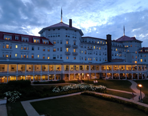 Historic hotel at dusk, featuring illuminated windows and a landscaped lawn with flowers, under a cloudy sky.