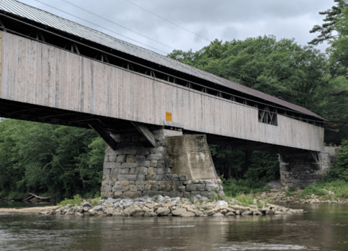 A wooden covered bridge spans a river, supported by stone pillars, surrounded by lush greenery and a cloudy sky.