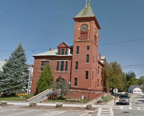 Historic red brick building with a clock tower, surrounded by trees and a sidewalk, located at a street intersection.