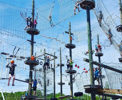 A vibrant ropes course with people climbing and navigating obstacles among tall wooden structures against a blue sky.