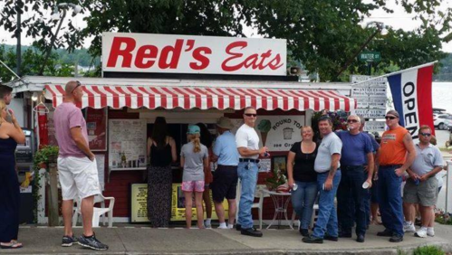 A busy outdoor eatery named "Red's Eats" with a striped awning, people waiting in line, and a nearby "OPEN" sign.