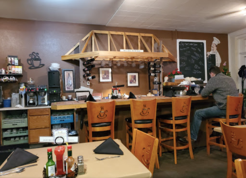 A cozy café interior with wooden bar stools, a counter, and a man sitting at the bar, surrounded by coffee equipment.