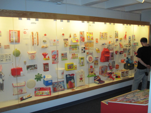 A display case filled with colorful toys and games from the 1970s, with a person observing the collection.