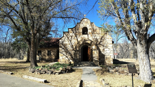 Historic stone chapel surrounded by trees, with a clear blue sky and a pathway leading to the entrance.