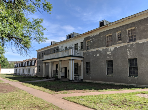 Abandoned building with peeling paint, a porch, and overgrown grass, set against a clear blue sky.