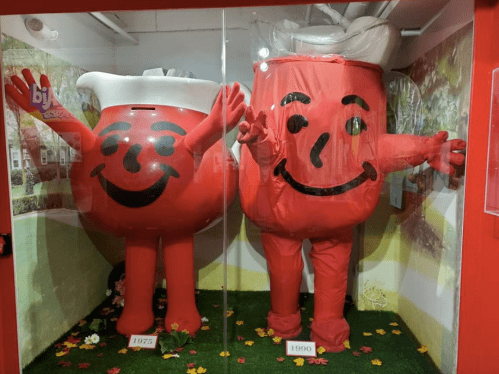 Two large, smiling Kool-Aid Man mascots from 1975 and 1990 displayed in a glass case with a colorful background.