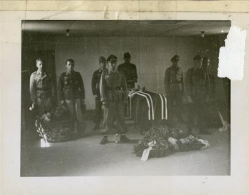 Black and white photo of soldiers standing around a casket with floral tributes in a dimly lit room.
