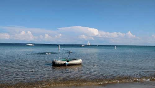 A child stands in shallow water near a small inflatable boat, with a sailboat and clouds in the background.