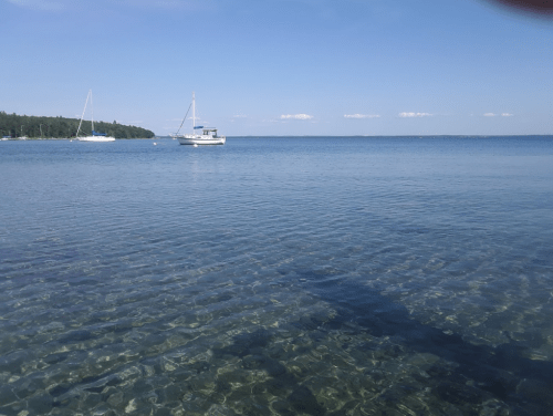 A calm, clear lake with a boat in the distance and sailboats anchored nearby, surrounded by trees under a blue sky.