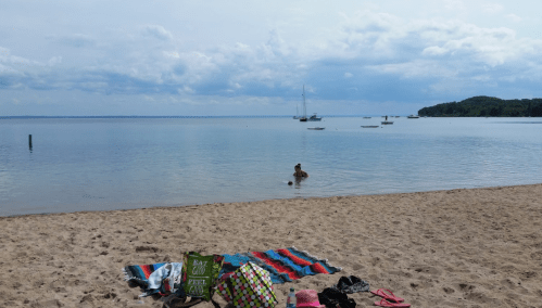 A sandy beach with a person wading in calm water, boats in the distance, and colorful towels on the shore.