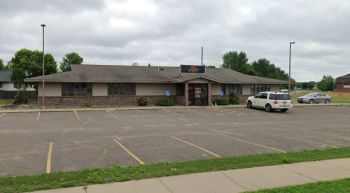 A single-story building with a parking lot, surrounded by greenery, under a cloudy sky.
