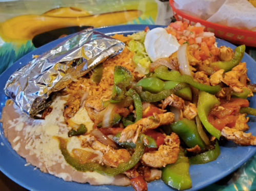 A colorful plate of chicken fajitas with bell peppers, rice, refried beans, lettuce, tomatoes, and a side of sour cream.