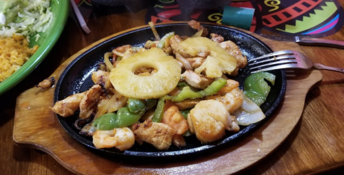 Sizzling fajitas with chicken, shrimp, bell peppers, and pineapple on a wooden platter, served with rice.