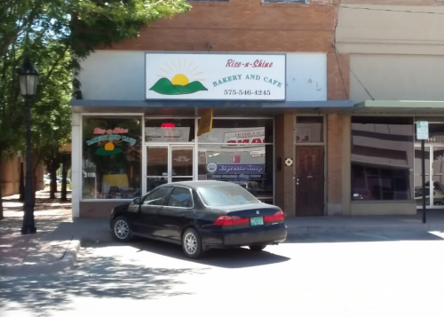 A bakery and café storefront with a sign reading "Rise-n-Shine Bakery and Café" and a parked black car outside.