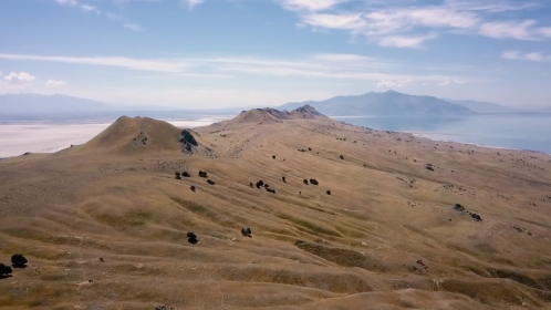 Aerial view of rolling hills and mountains under a blue sky, with a body of water in the distance.
