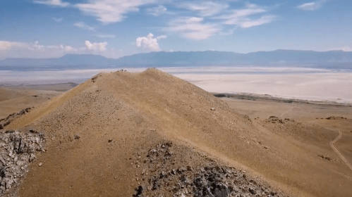Aerial view of a sandy hill with a vast salt flat and mountains in the background under a blue sky with clouds.