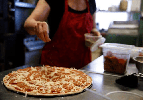 A person in an apron sprinkles seasoning on a pizza topped with sauce and cheese in a kitchen setting.