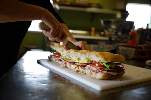 A person slices a large sandwich filled with meats, lettuce, and tomatoes on a cutting board in a kitchen.